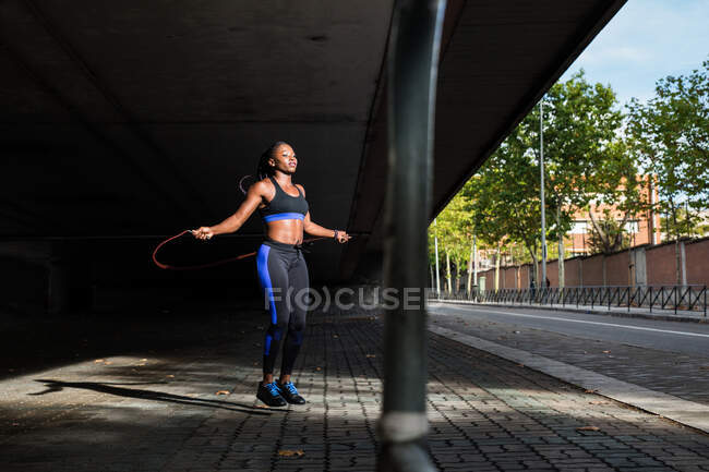 Strong African American female in sportswear jumping with skipping rope while exercising on city street on sunny day — Stock Photo