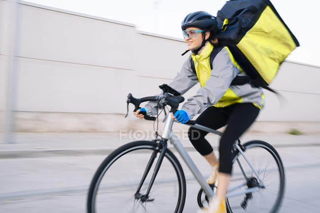 Happy female courier with thermal bag riding bike on street road while delivering food in city, motion blur — Stock Photo