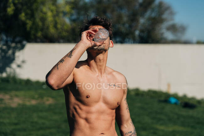 Atleta masculino com tatuagem bebendo água de garrafa após o treinamento em luz solar — Fotografia de Stock
