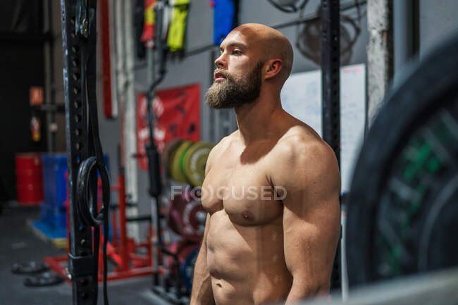 Uomo muscoloso barbuto guardando lontano mentre in piedi vicino alle attrezzature durante l'allenamento in palestra moderna — Foto stock