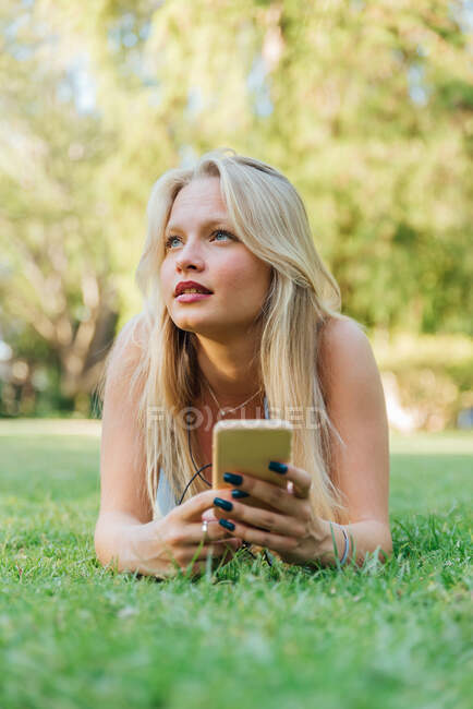 Charming female lying on grass in park and listening to music in headphones in summer — Stock Photo