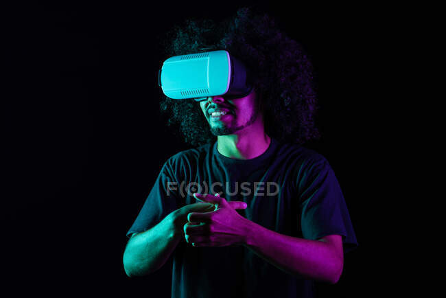 Homme latin excité avec coiffure afro et lunettes VR expérimentant la réalité virtuelle sur fond noir en studio avec néons — Photo de stock