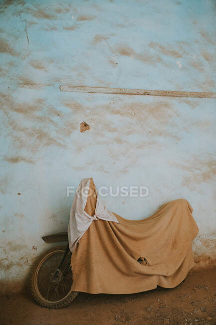 Motocicleta cubierta con tela y aparcada cerca de la pared erosionada en la calle de Marrakech, Marruecos - foto de stock
