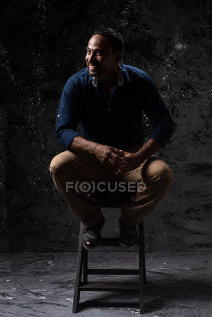 Delighted stylish ethnic male sitting on wooden stool in studio on dark background and looking away — Stock Photo