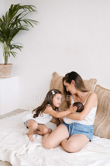 Нежная маленькая девочка трогает новорожденного, сидя на полу с матерью дома — стоковое фото