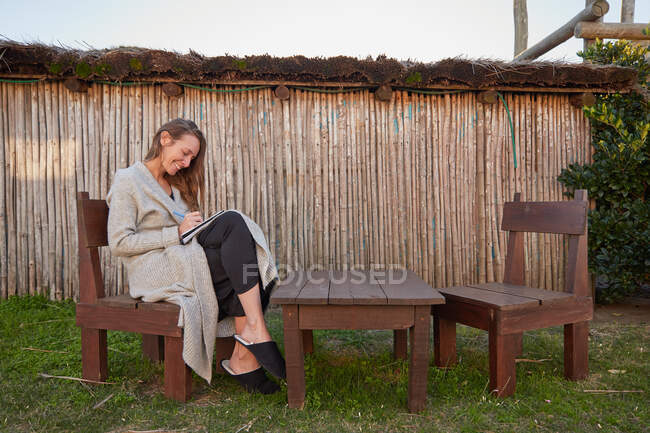 Боковой вид восхищенной женщины, пишущей в тетрадке, сидя на деревянном стуле на заднем дворе летом — стоковое фото
