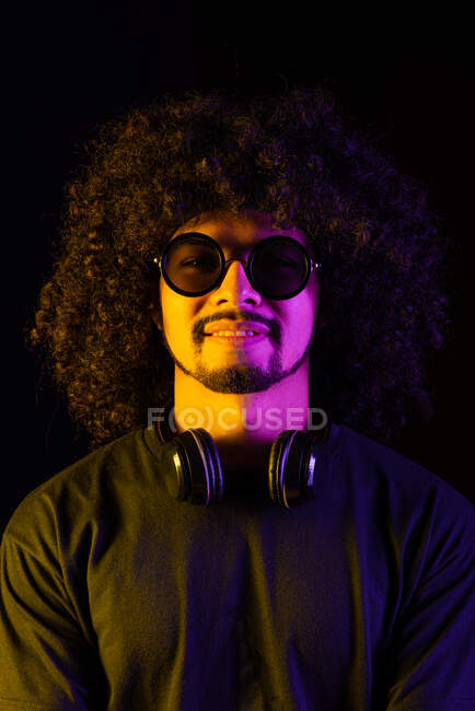 Positive curly haired Latin male in stylish sunglasses and with headphones on neck looking at camera on black background in studio with yellow and pink neon lights — Stock Photo