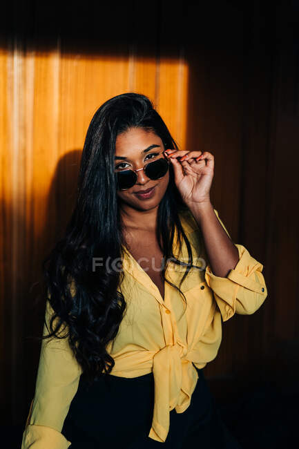 Jeune femme hispanique en chemise jaune tendance regardant la caméra et ajustant les lunettes de soleil tout en se tenant dans l'ombre — Photo de stock