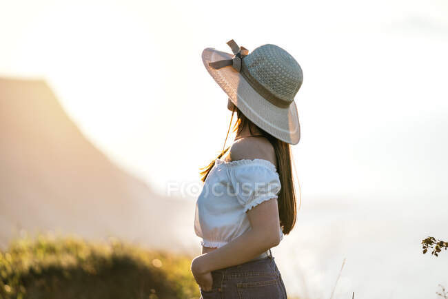 Vista lateral de la mujer soñadora anónima en jeans y top blanco y mirando hacia otro lado en pensamientos agradables mientras está de pie en un exuberante terreno cubierto de hierba en un día soleado - foto de stock