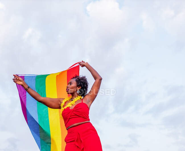 De baixo da mulher afro-americana elegante no desgaste da moda levantando bandeira com ornamento do arco-íris, enquanto olha para longe na estrada — Fotografia de Stock