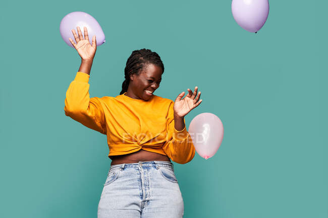 Возбужденная афроамериканка в джинсах и желтом топе стоящая рядом с падающими воздушными шарами на синем фоне в студии — стоковое фото