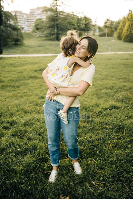 Усміхнена молода мати носить милу маленьку доньку в літньому одязі з браслетом і дивиться геть, стоячи на зеленому газоні в літньому парку — стокове фото