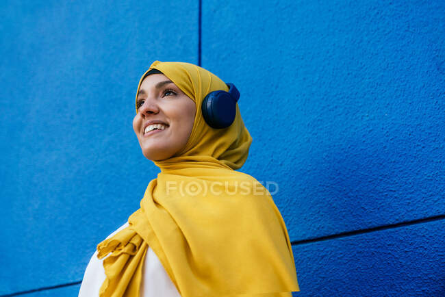 Cheerful muslim female in hijab listening to music in headphones on blue background in city — Stock Photo