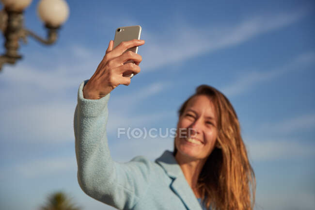 Low angle of smiling adult lady in warm coat taking selfie on phone under blue cloudy sky in city street in sunny day — Stock Photo