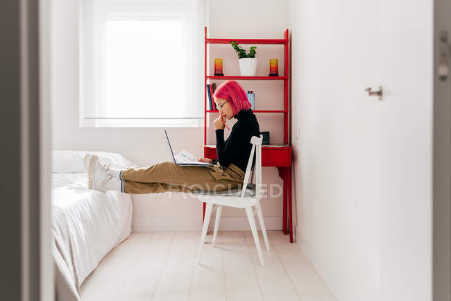 Vista laterale di focalizzata giovane donna freelance in abiti casual seduto sulla sedia e utilizzando il computer portatile mentre si lavora sul progetto in leggero appartamento moderno — Foto stock
