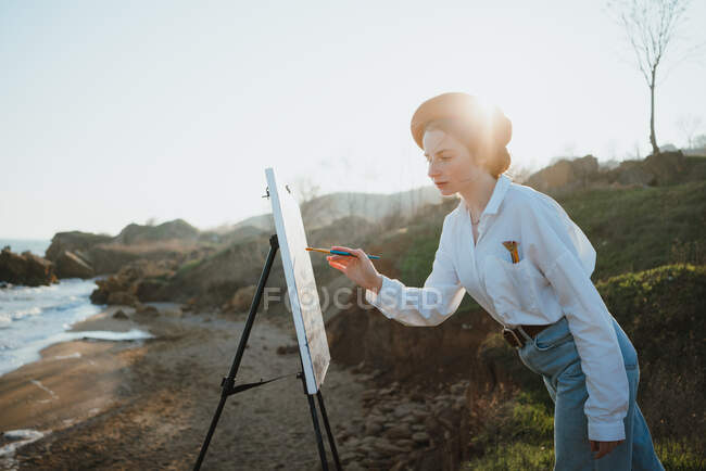 Вид збоку молодої жінки в стильному одязі і береті, що стоїть на трав'яному узбережжі біля піску і океану в сонячний день, малюючи картину пензлем на мольберті — стокове фото
