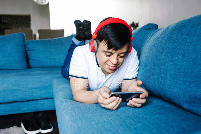 Delighted focus Latin teen boy in headphones playing videogame on mobile phone while lying on sofa at home — Stock Photo