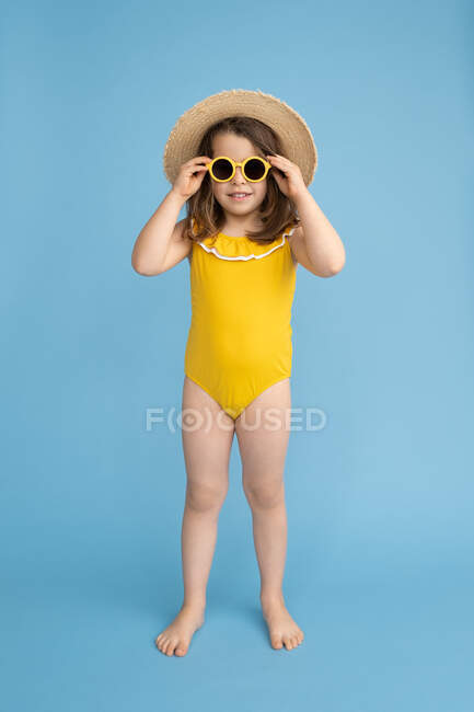 Полное тело милой счастливой маленькой девочки в желтом купальнике и соломенной шляпе со стильными солнцезащитными очками, стоящими на синем фоне и смотрящими в камеру — стоковое фото