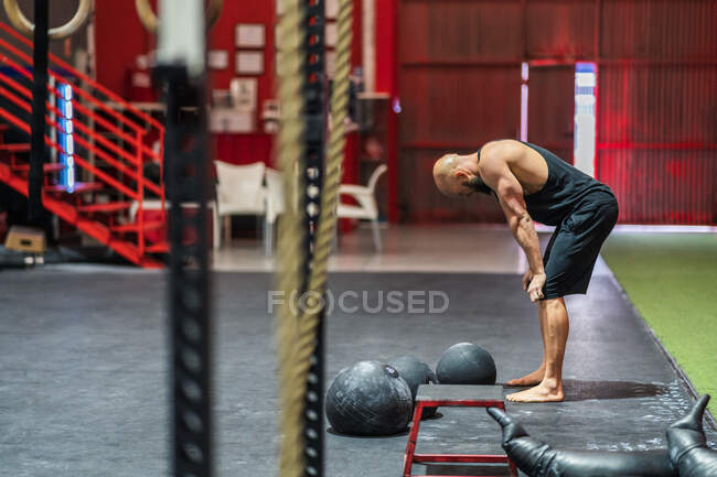 Vista lateral do homem careca muscular em sportswear inclinando-se sobre os joelhos e descansando durante o treinamento funcional em ginásio espaçoso — Fotografia de Stock