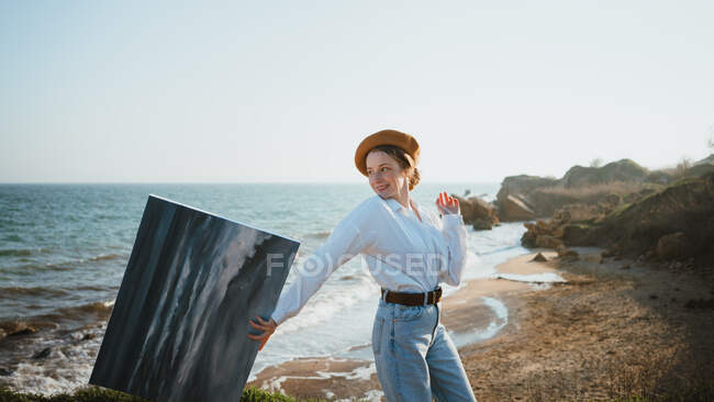 Вид сбоку позитивной молодой художницы в стильном наряде и шляпе, прогуливающейся возле песчаного пляжа волнистого моря с картиной в руке — стоковое фото
