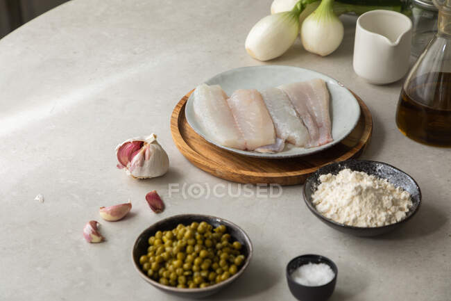 Dall'alto spicchi d'aglio freschi e sale posto sul tavolo vicino al filetto di nasello, ciotola con piselli e farina durante la preparazione del cibo in cucina — Foto stock