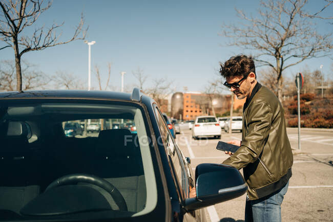 Side view of male in leather jacket and sunglasses with cellphone opening automobile door on pavement — Stock Photo