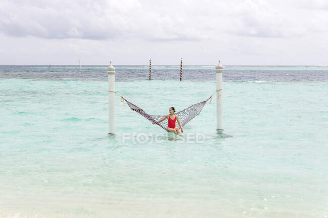 Frau in rotem Badeanzug sitzt in Hängematte und schwingt sich an bewölkten Tagen über die Surfleine des Meeres und entspannt sich auf den Malediven — Stockfoto
