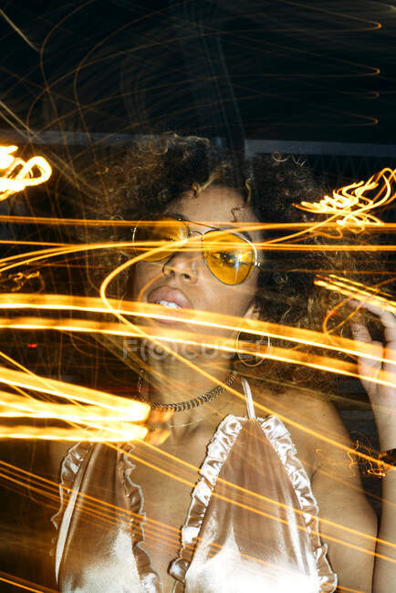 Femme afro-américaine auto-assurée avec des cheveux bouclés dans des lunettes de soleil à la mode et haut relaxant dans la rue le soir près de lumières gelées — Photo de stock