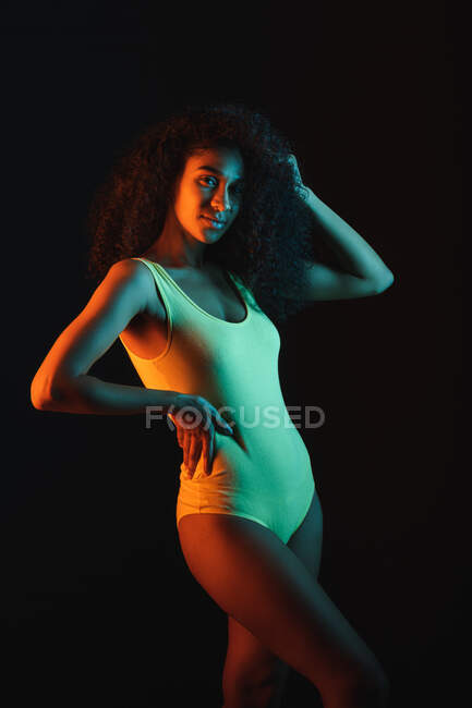 Joven mujer afroamericana en ropa interior con la mano en la cintura tocando el pelo rizado mientras mira la cámara - foto de stock
