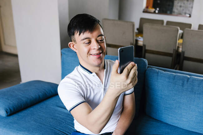 Ragazzo latino sorridente con sindrome di Down che si spara da solo sullo smartphone mentre si siede sul divano a casa — Foto stock