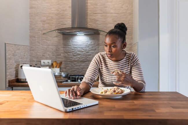 Afroamericano femmina godendo gustoso patacon con topping durante la navigazione internet su netbook in cucina a casa — Foto stock