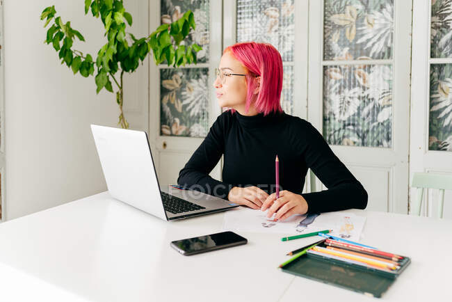 Молодая креативная женщина-дизайнер в повседневной одежде и очках задумчиво смотрит в сторону, сидя за столом с гаджетами и рисуя эскизы с красочными карандашами — стоковое фото