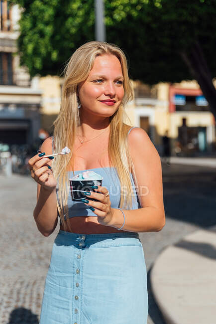 Belle blonde jeune femme mangeant froid savoureux crème glacée tout en se tenant dans la rue de la ville par une journée ensoleillée en été — Photo de stock