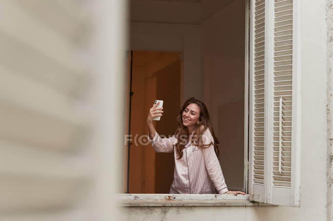 Жінка в піжамі, що стоїть біля вікна і приймає мобільний телефон селфі вдома — стокове фото