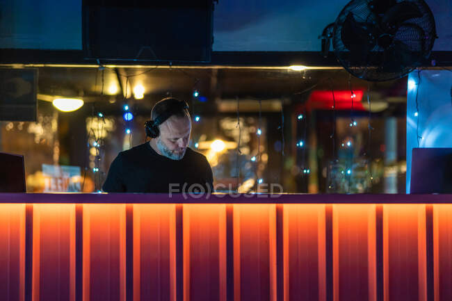 Bajo ángulo de hombre serio de mediana edad en auriculares inalámbricos que se concentran y juegan controlador DJ mientras se realiza en concierto en el vibrante mostrador naranja en el escenario en la discoteca moderna - foto de stock