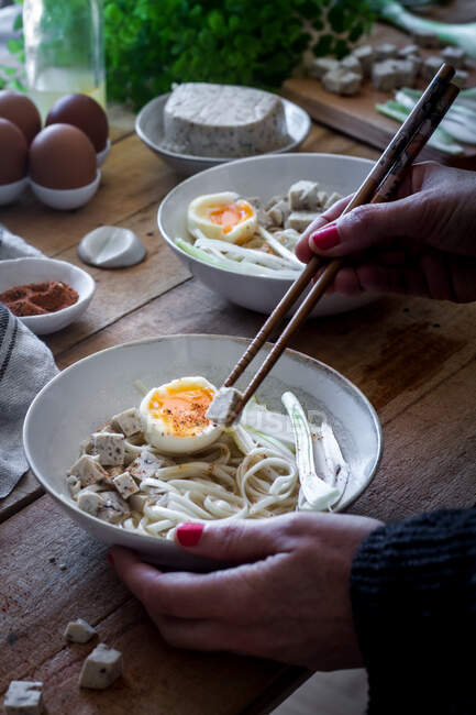 Cropped unrecognizable person preparing fresh cooked ramen noodles with tofu, eggs and vegetables with chopsticks on a wooden table — Stock Photo