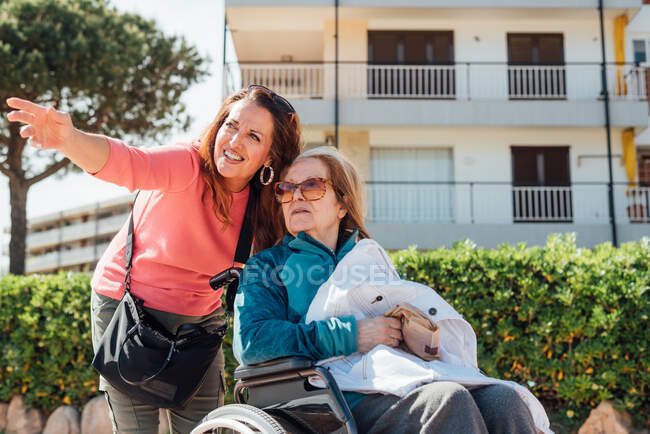 Happy adult woman pushing wheelchair with senior mother while walking on the street in city during summer — Stock Photo