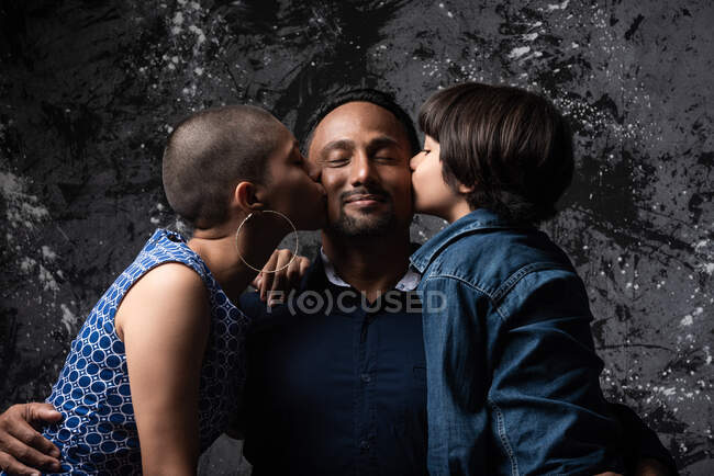 Multiethnic loving woman and teenage son kissing man on cheek on dark background in studio — Stock Photo
