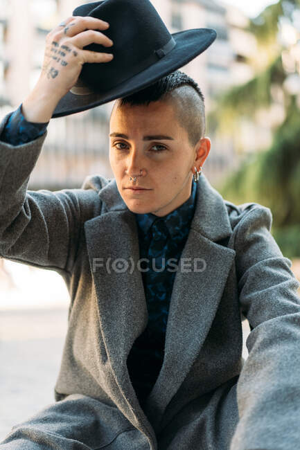 Queer in stylischer Kleidung und Hut mit Tätowierung und Mohawk blickt in die Kamera, während er in der Stadt sitzt — Stockfoto