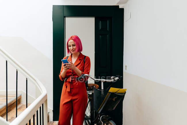 Junge stylische Frau im roten Anzug mit Rucksack und Smartphone, während sie mit Fahrrad im Treppenhaus steht — Stockfoto