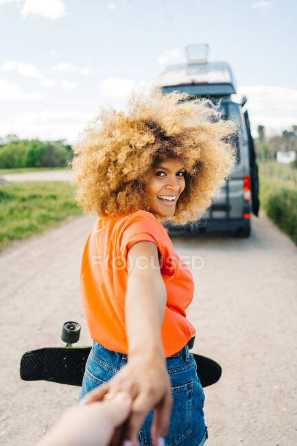 Feliz mujer afroamericana con longboard sonriendo y mirando a la cámara mientras conduce amigo de la cosecha a la camioneta durante el viaje por carretera - foto de stock