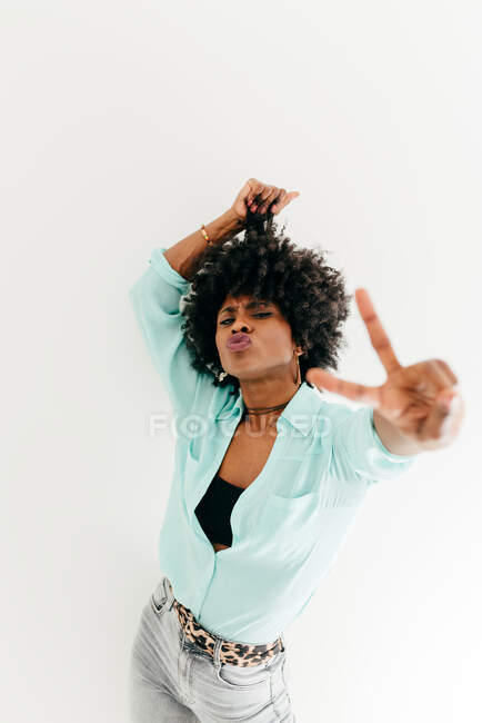 Playful young African American female in trendy outfit having fun and showing peace sign on white background — Stock Photo