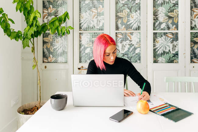 Diseñadora freelance femenina pensativa en atuendo casual y gafas trabajando en la mesa con portátil y dibujando bocetos de moda con lápices - foto de stock