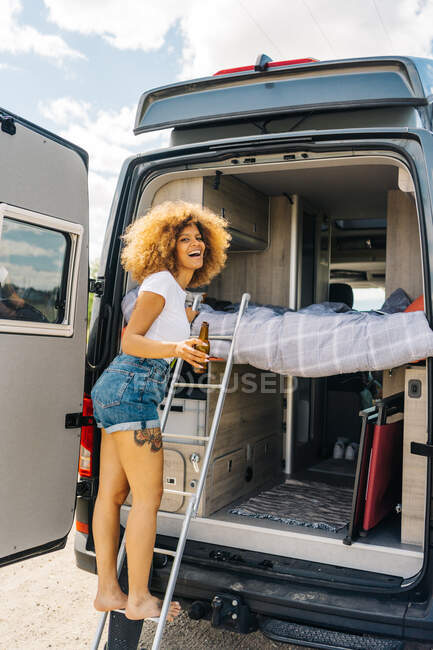 Vue latérale d'une femme noire enchantée avec de la bière riant et regardant la caméra debout sur l'échelle à l'extérieur du camping-car et grimpant sur le lit le jour d'été dans la campagne — Photo de stock