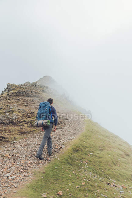 Vue arrière du randonneur masculin sans visage marchant le long du sentier dans les hautes terres pendant la randonnée par temps brumeux au Pays de Galles — Photo de stock