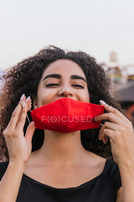 Positivo giovane dai capelli ricci femmina ispanica togliersi il tessuto rosso maschera protettiva per la prevenzione coronavirus mentre trascorre la giornata estiva in strada — Foto stock