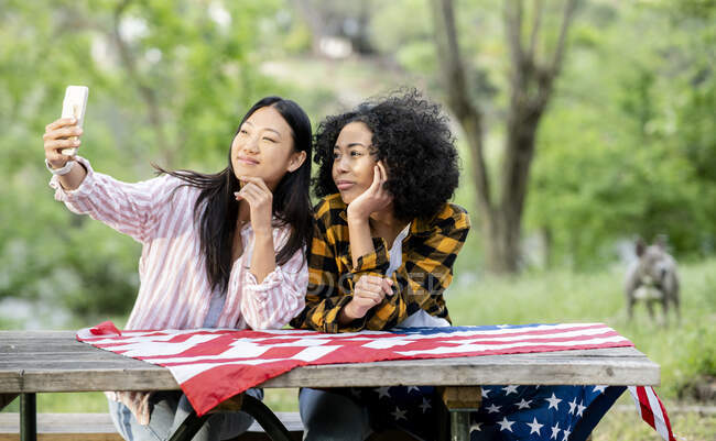Fröhliches Paar multiethnischer lesbischer Frauen, die mit amerikanischer Flagge im Wald am Tisch sitzen und gemeinsam Selbstaufnahmen mit dem Handy machen — Stockfoto