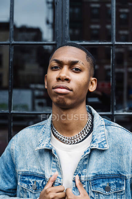 Black trendy serious male with silver chain on neck in blue denim jacket looking at camera on street — Stock Photo