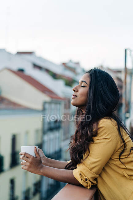 Vista lateral da jovem mulher hispânica com olhos fechados desfrutando de bebida quente enquanto descansa na varanda à noite — Fotografia de Stock