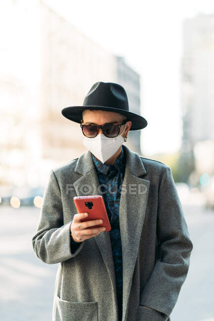 Квір у стильному верхньому одязі та текстових повідомленнях респіраторної маски на мобільному телефоні в місті під час пандемії COVID 19 — стокове фото
